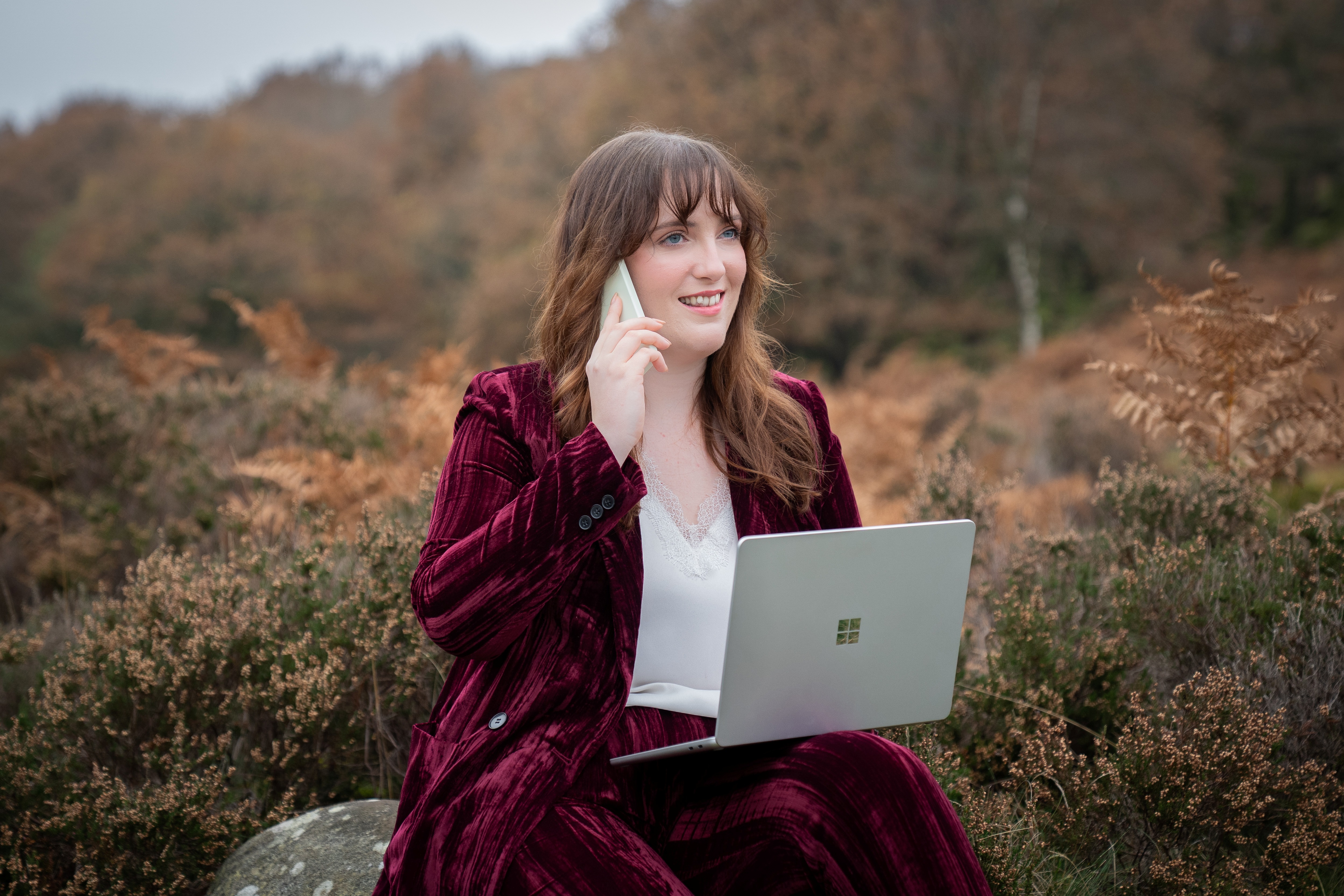 Nord-Essence founder Rhiannon sits on a rock in an autumn woodland, wearing a burgundy velvet suit and white blouse. She holds a silver laptop and speaks on a phone, surrounded by fallen leaves and fading greenery — blending business with nature’s quiet.