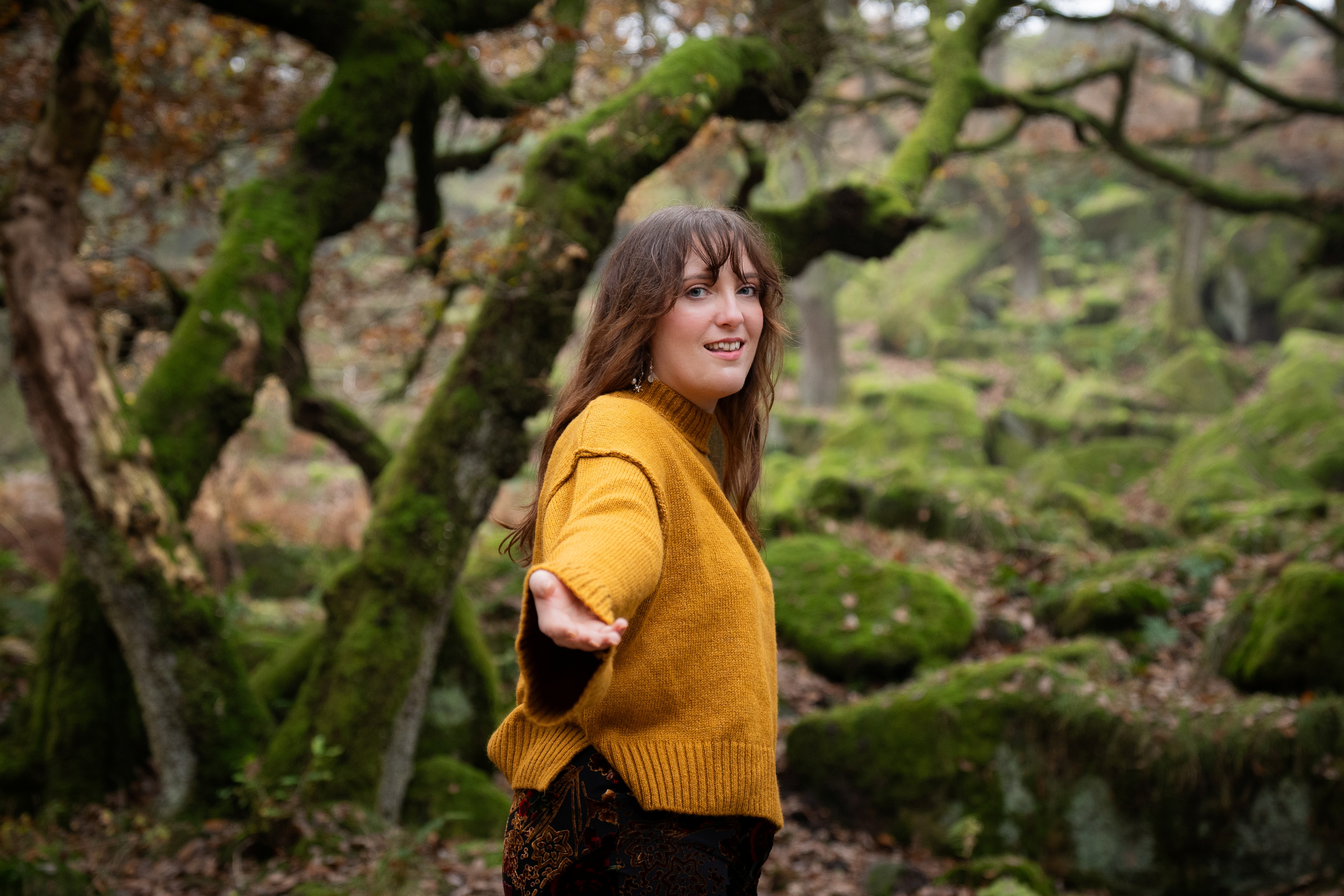 Nord-Essence founder Rhiannon in a mustard sweater stands in a moss-covered forest, arm extended in invitation. Twisted tree trunks and vibrant green moss surround her, creating a sense of mystery, connection, and quiet guidance through nature.