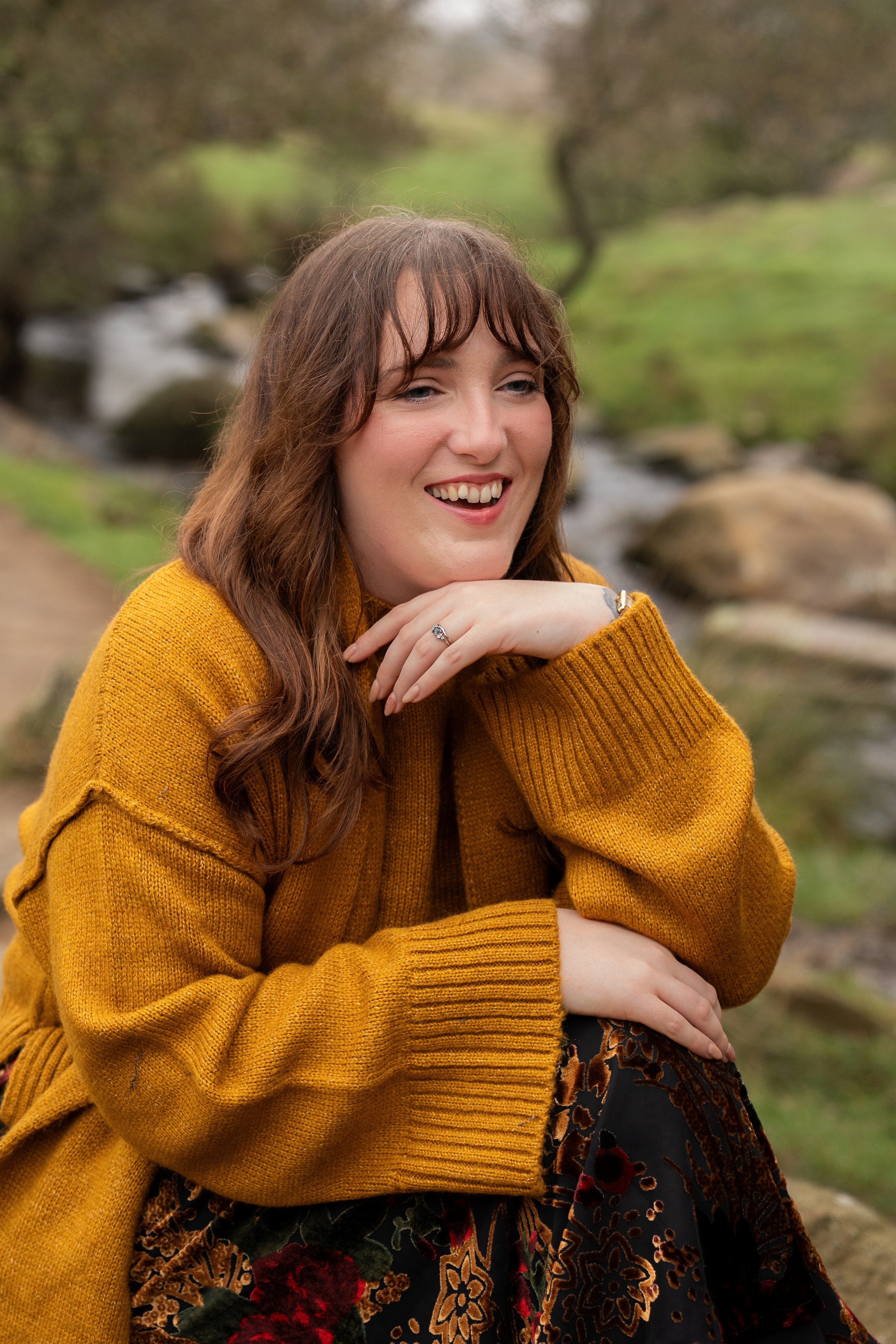 Nord-Essence Founder Rhiannon A woman sits near a stream in a forest clearing, wearing a mustard yellow sweater and a floral-patterned skirt. Her long, wavy brown hair frames a warm smile as she rests one arm on her knee — a joyful, grounded moment in nature