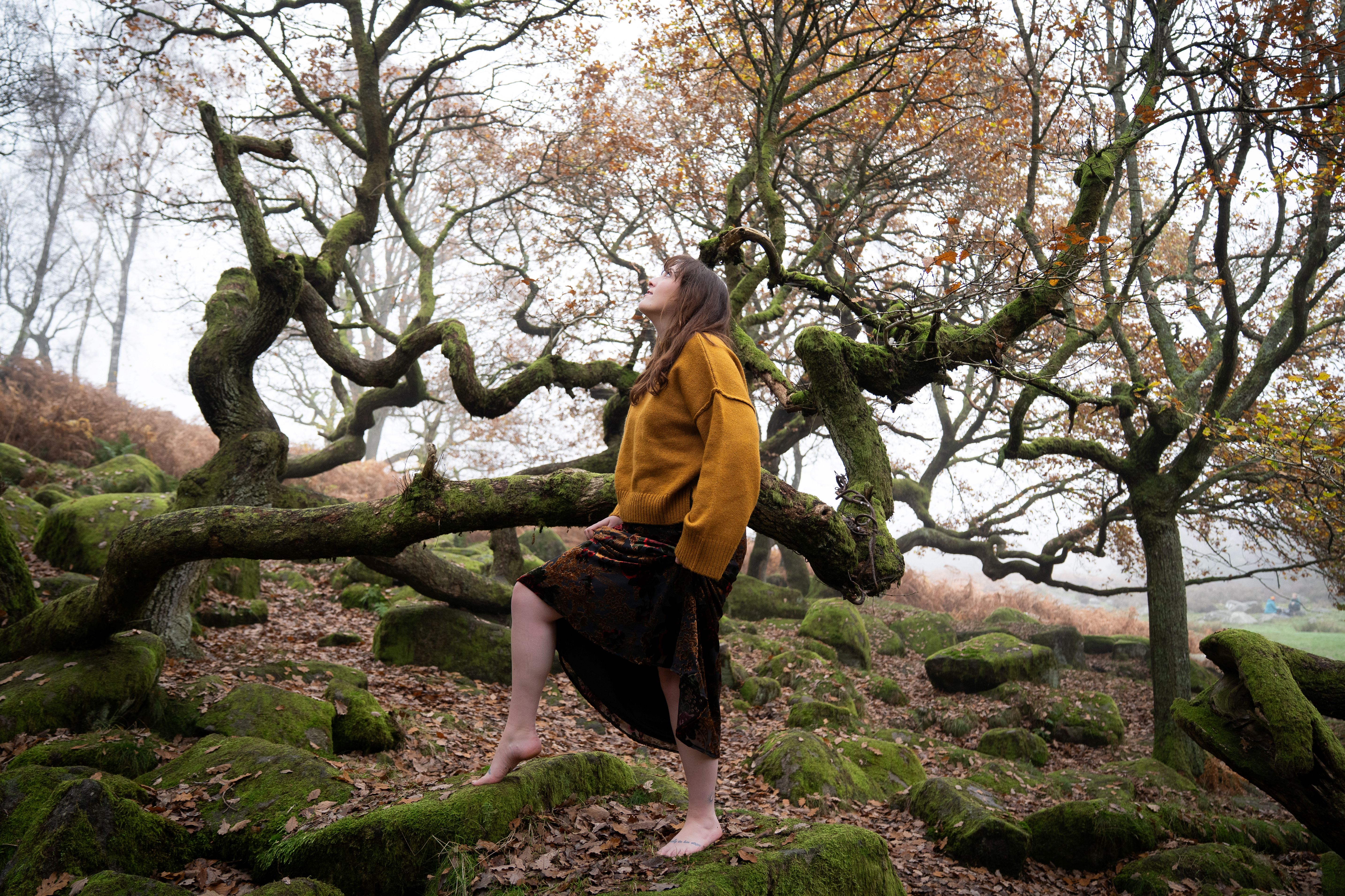 Nord-Essence founder Rhiannon stands barefoot on a moss-covered rock in a misty forest, wearing a mustard yellow sweater and dark skirt. One leg is raised onto another rock as she gazes upward, surrounded by twisted leafless trees and fallen leaves — a moment of grounded strength and quiet connection with nature.