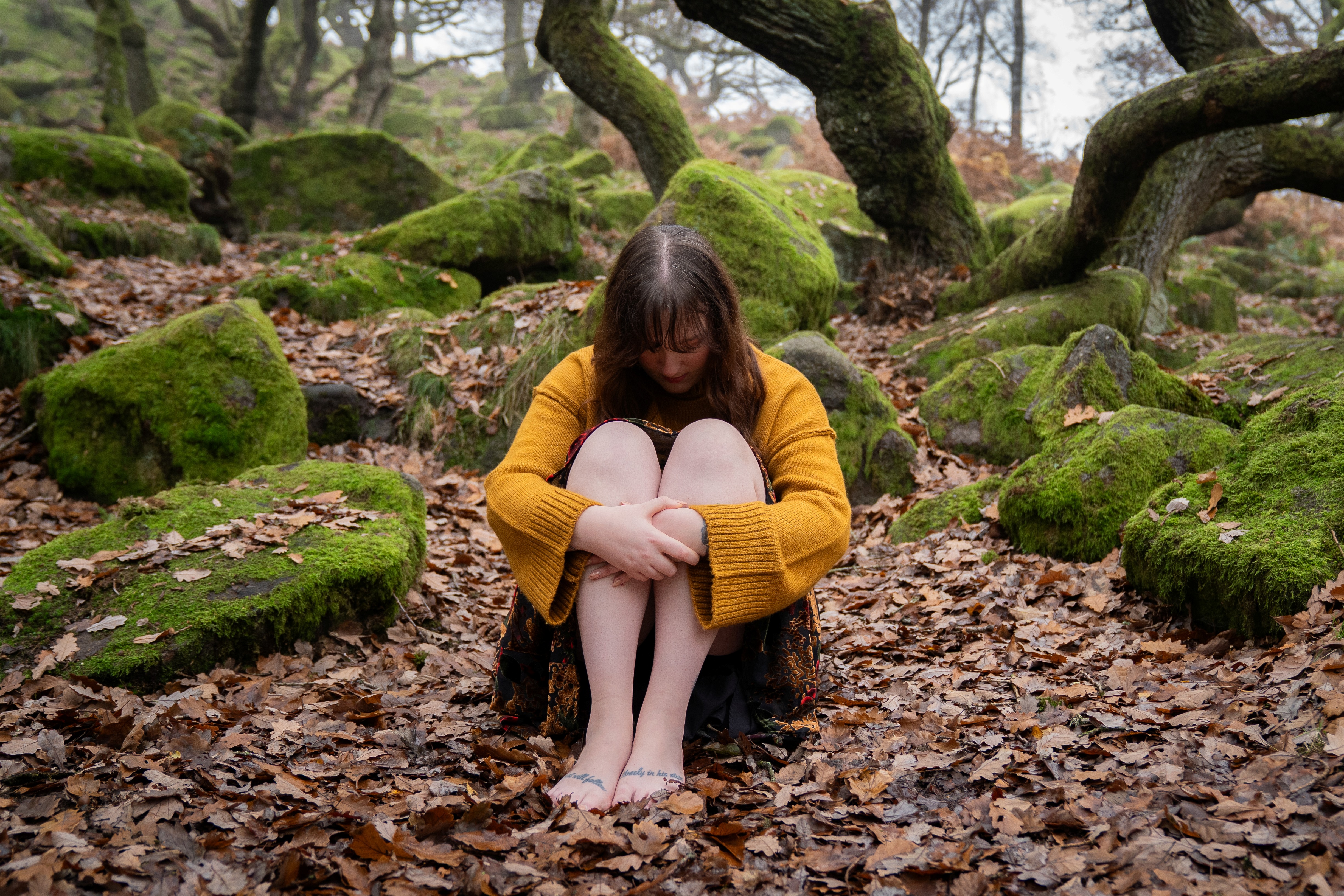 Nord-Essence founder Rhiannonsits barefoot on a forest floor covered in dry leaves, wearing a mustard yellow sweater and patterned skirt. Her arms wrap around her knees and her head is lowered, surrounded by mossy rocks and twisted tree trunks — a quiet moment of introspection and emotional depth.