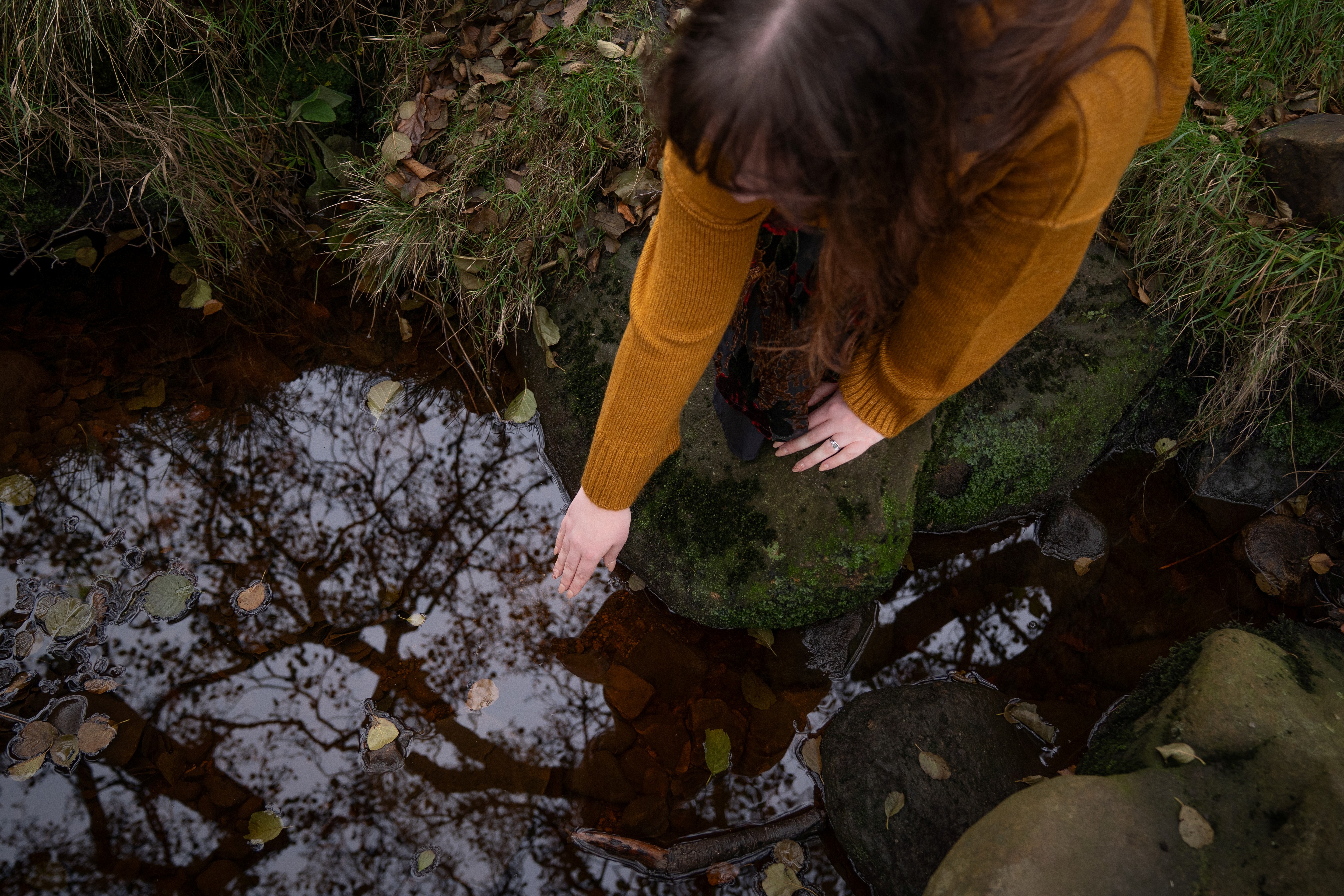 Nord-Essence founder Rhiannon in a mustard sweater reaches toward a still stream, noticing her reflection in the water standing on mossy rocks surrounded by fallen leaves. Tree branches reflect in the water’s surface, creating a quiet moment of connection with nature