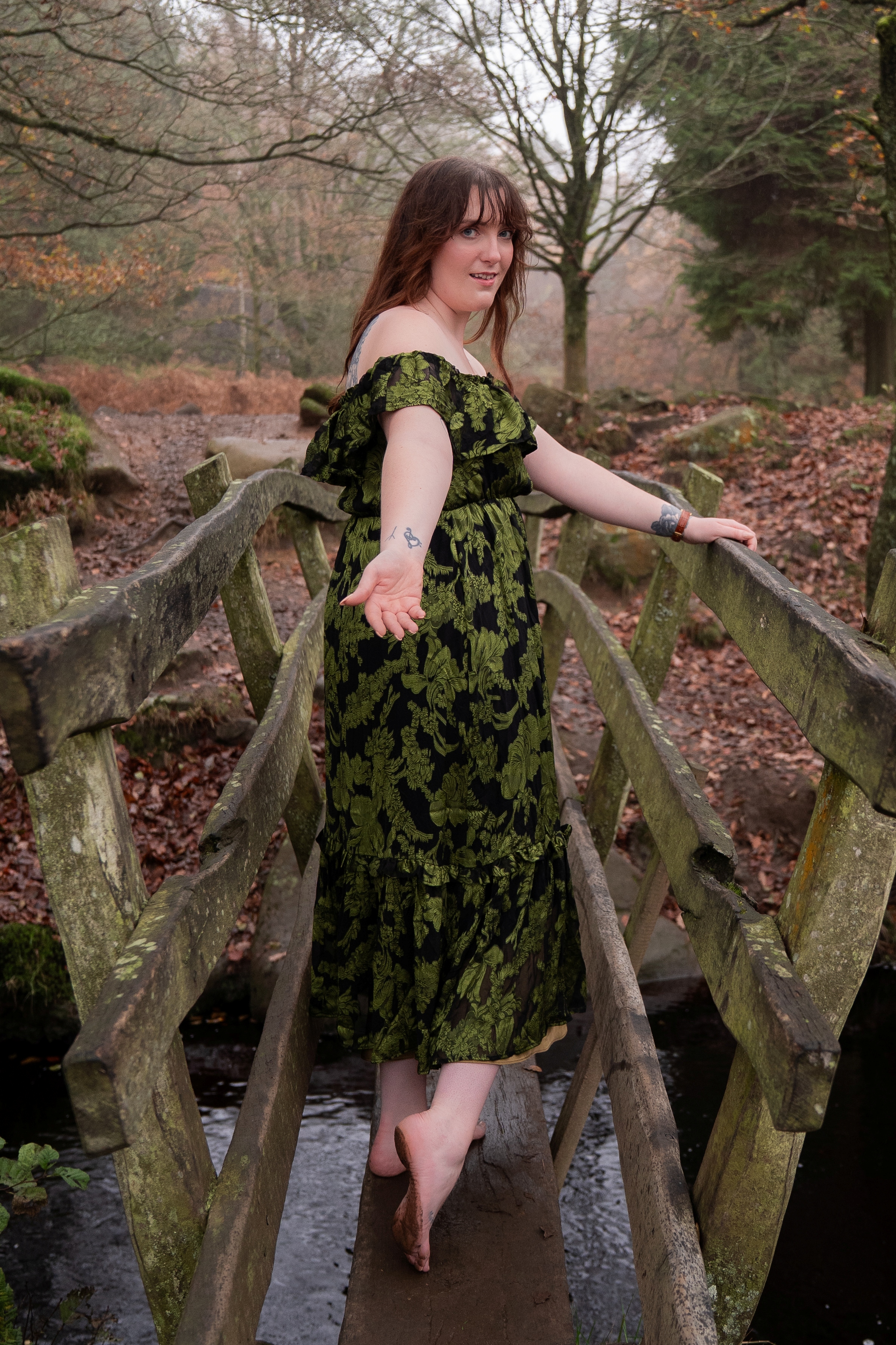 Nord-Essence founder Rhiannon stands barefoot on a rustic wooden bridge in a misty autumn forest, wearing an off-the-shoulder green and black patterned dress. She extends one arm behind her, palm up, and looks over her shoulder toward the camera — a quiet moment of embodied presence and rewilding.