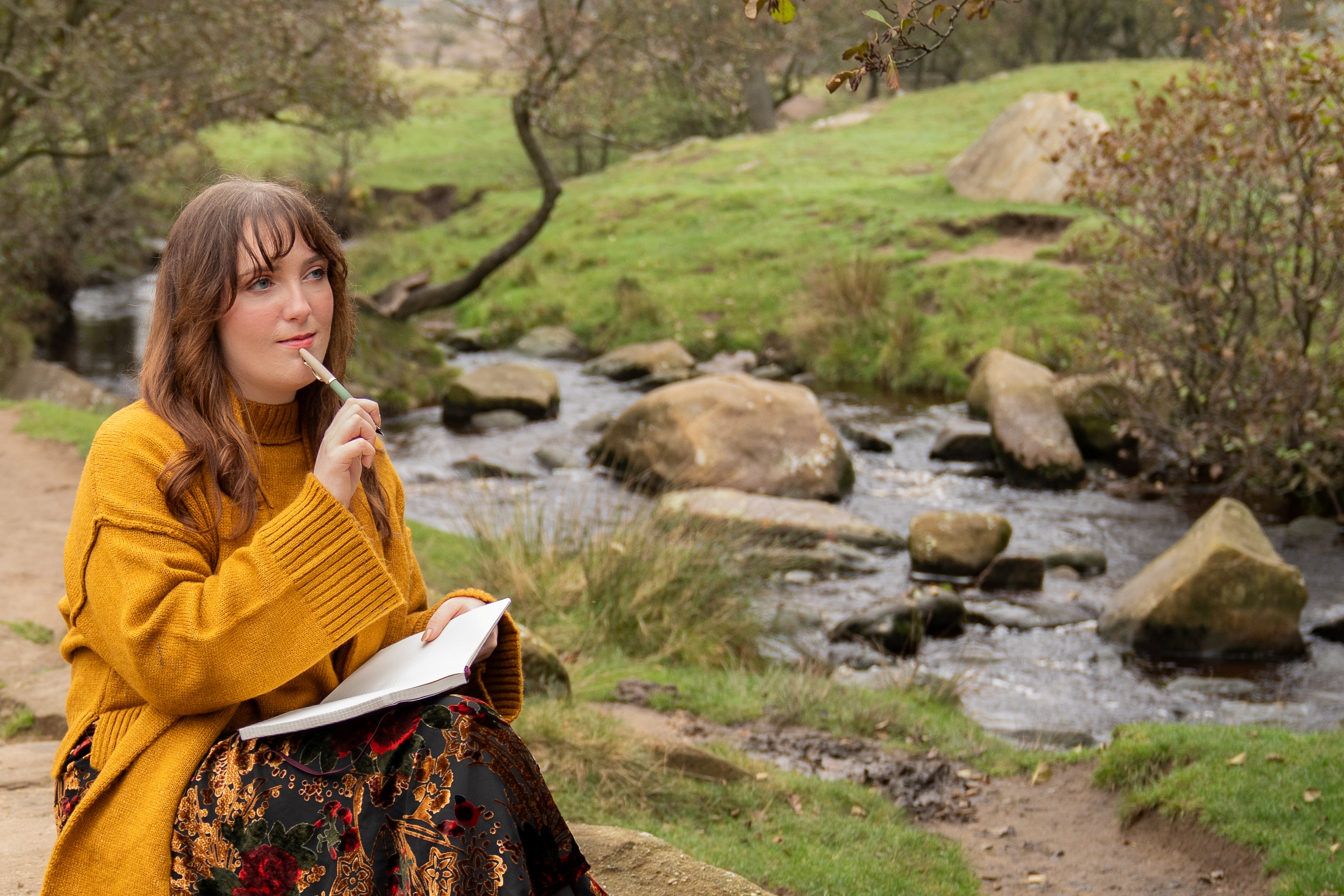Nord-Essence founder Rhiannon sits near a stream in an autumn forest, wearing a mustard yellow sweater and a long floral skirt. She holds a notebook and pen in a quiet moment of reflection, surrounded by rocks, trees, and fallen leaves.in a quiet, open field wearing a long dark dress with white abstract patterns. The landscape is softly lit, with bare trees, scattered leaves, and gentle hills suggesting late autumn. A large curved tree branch lies in the foreground, adding organic texture. The sky is partly cloudy with hints of blue, creating a peaceful, reflective atmosphere