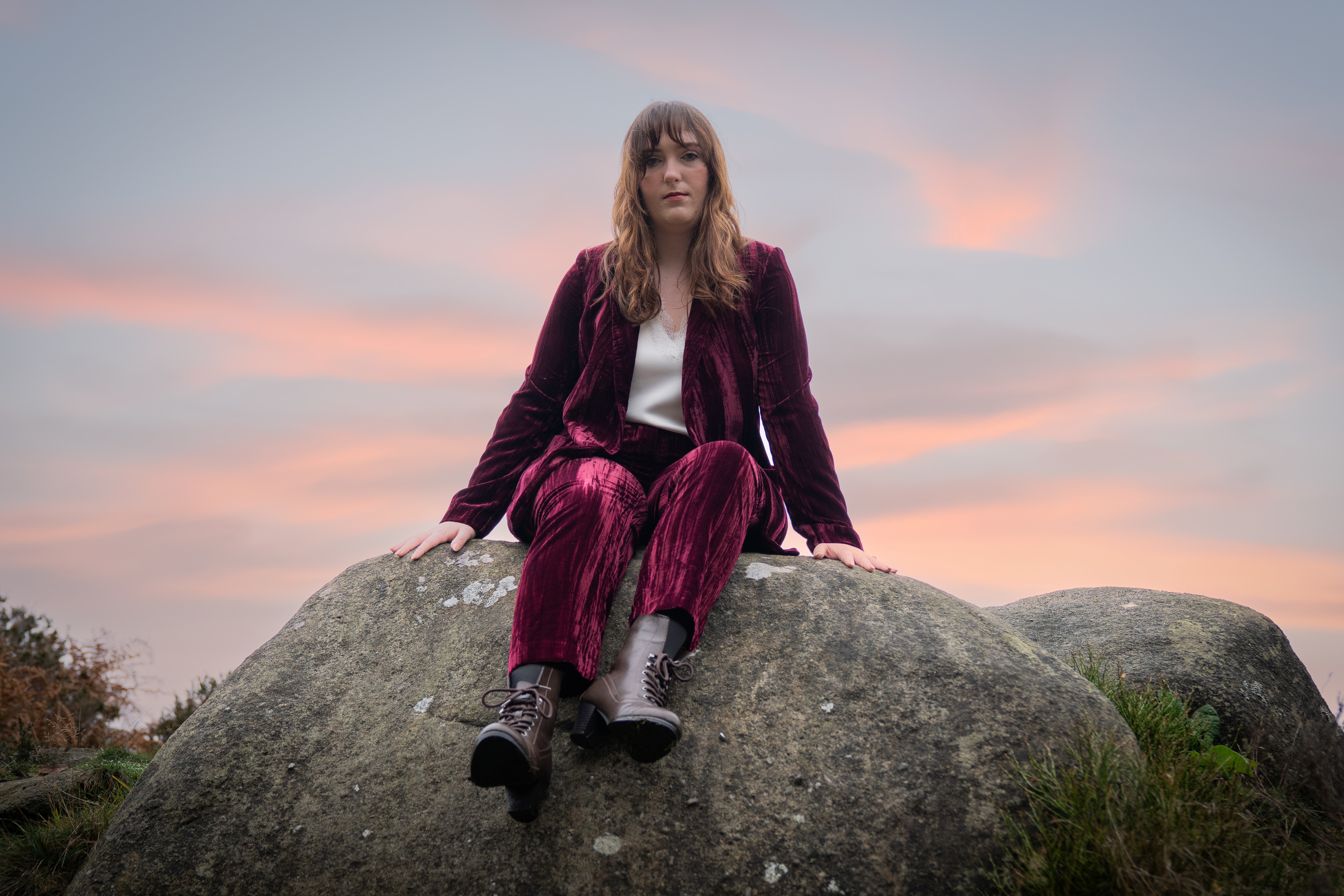 Nord-Essence founder Rhiannon A woman sits on a large rock at sunset, dressed in a deep red velvet suit and lace-up boots. The sky glows with soft pink and purple hues, casting a serene light over the grassy landscape — a contemplative moment of stillness and strength.