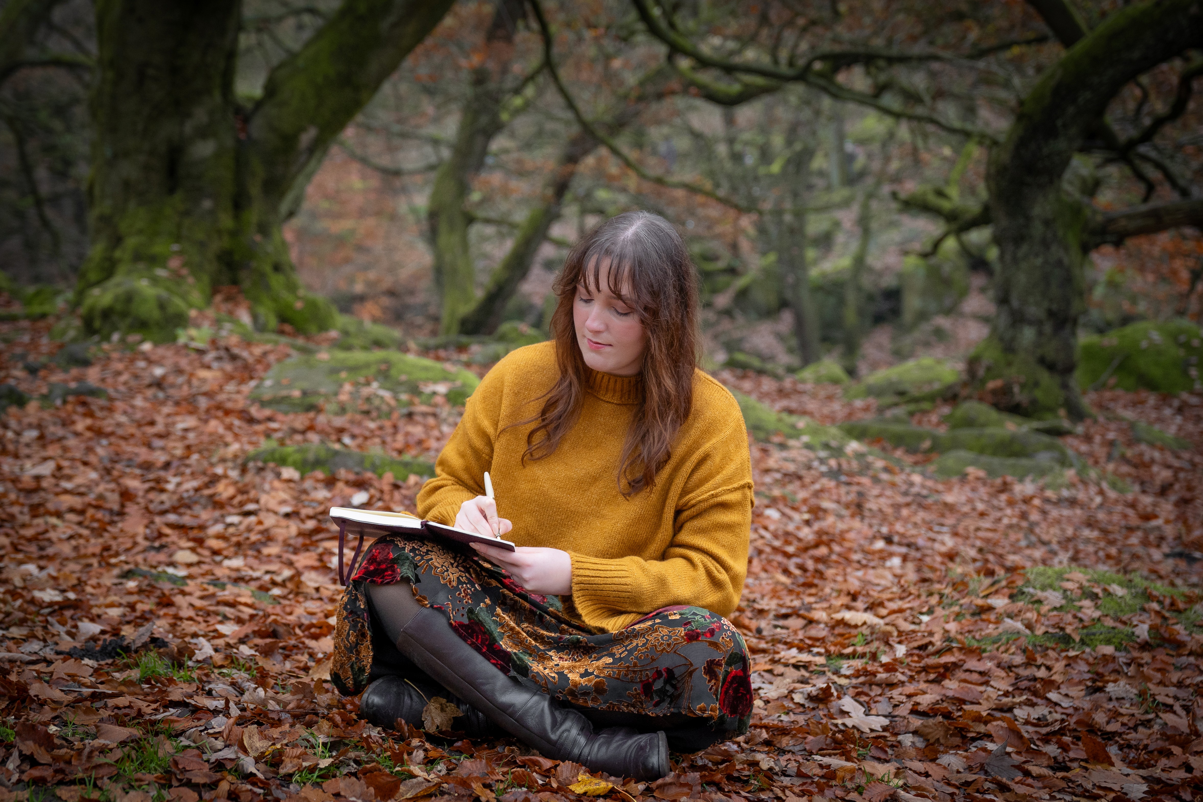 Nord-Essence founder Rhiannon sits cross-legged on a forest floor blanketed with autumn leaves, wearing a mustard yellow sweater and patterned skirt. She writes in a notebook, surrounded by mossy rocks and sparse trees — a quiet moment of creativity and grounded reflection.