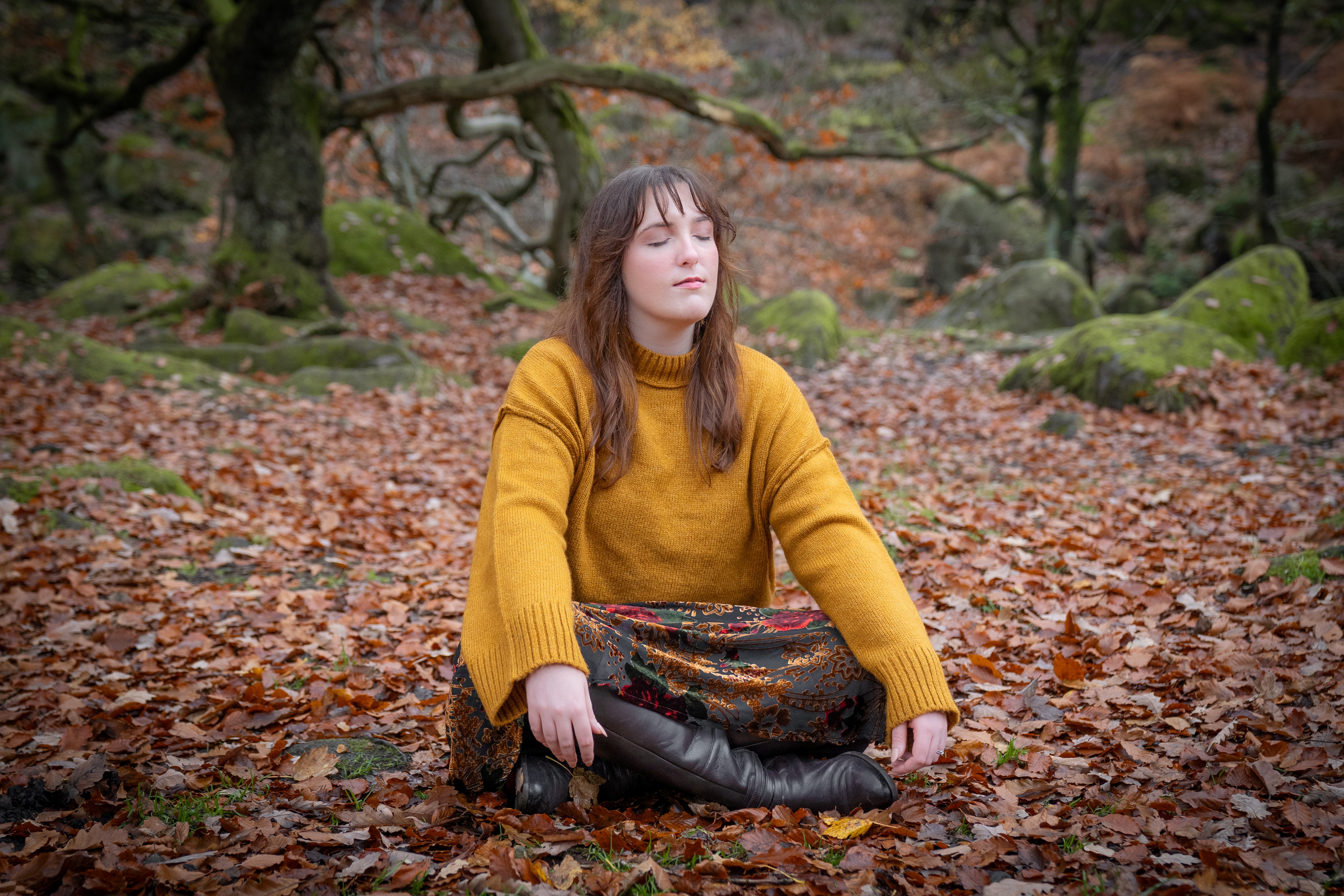Nord-Essence founder Rhiannon sits cross‑legged on a forest floor blanketed with autumn leaves, eyes closed in quiet reflection. She wears a mustard sweater and floral skirt, surrounded by mossy rocks and twisted branches — a moment of grounded solitude.